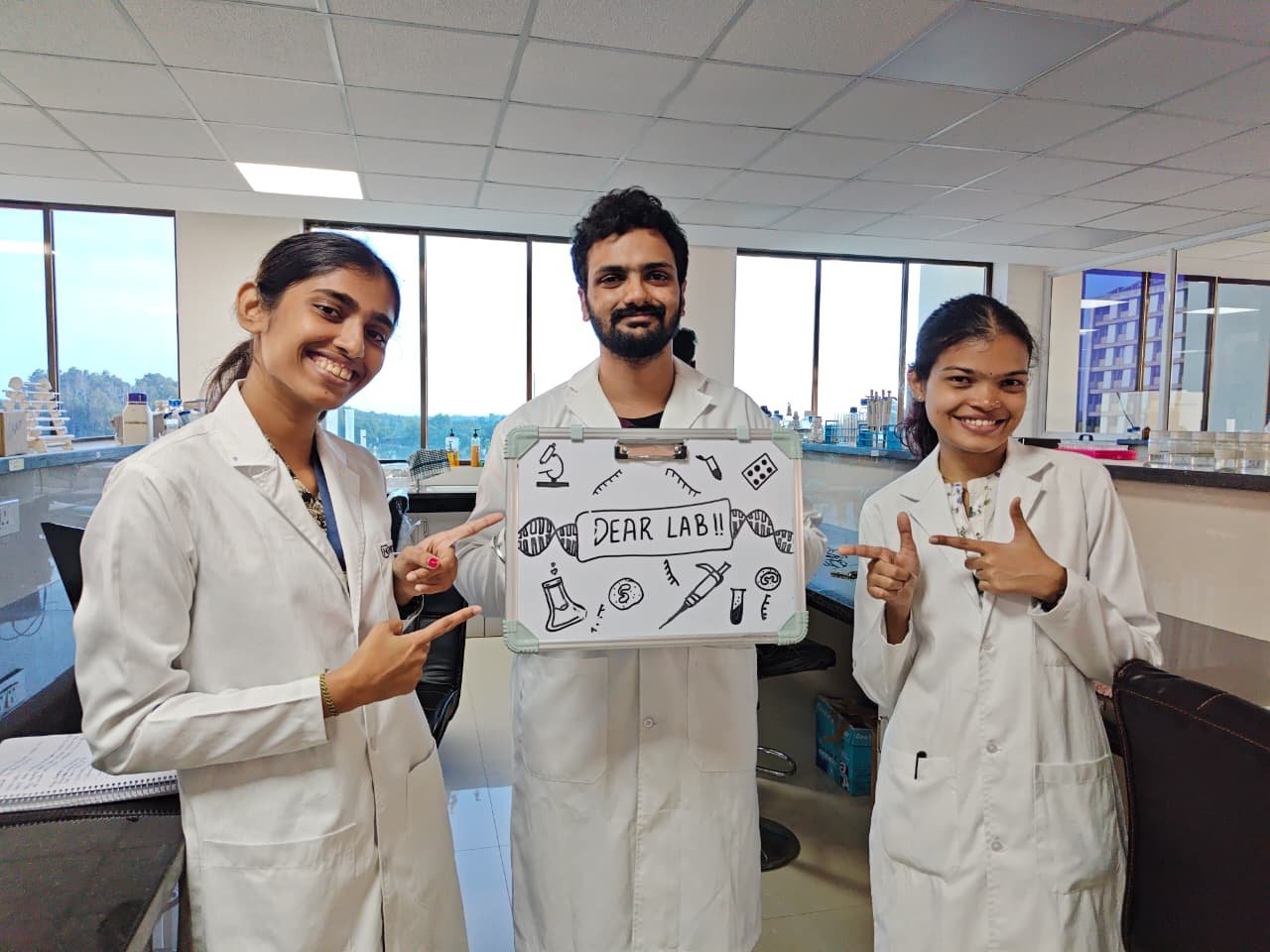 DEAR Lab members in lab coats holding a hand-drawn &ldquo;DEAR LAB!!&rdquo; sign in the lab